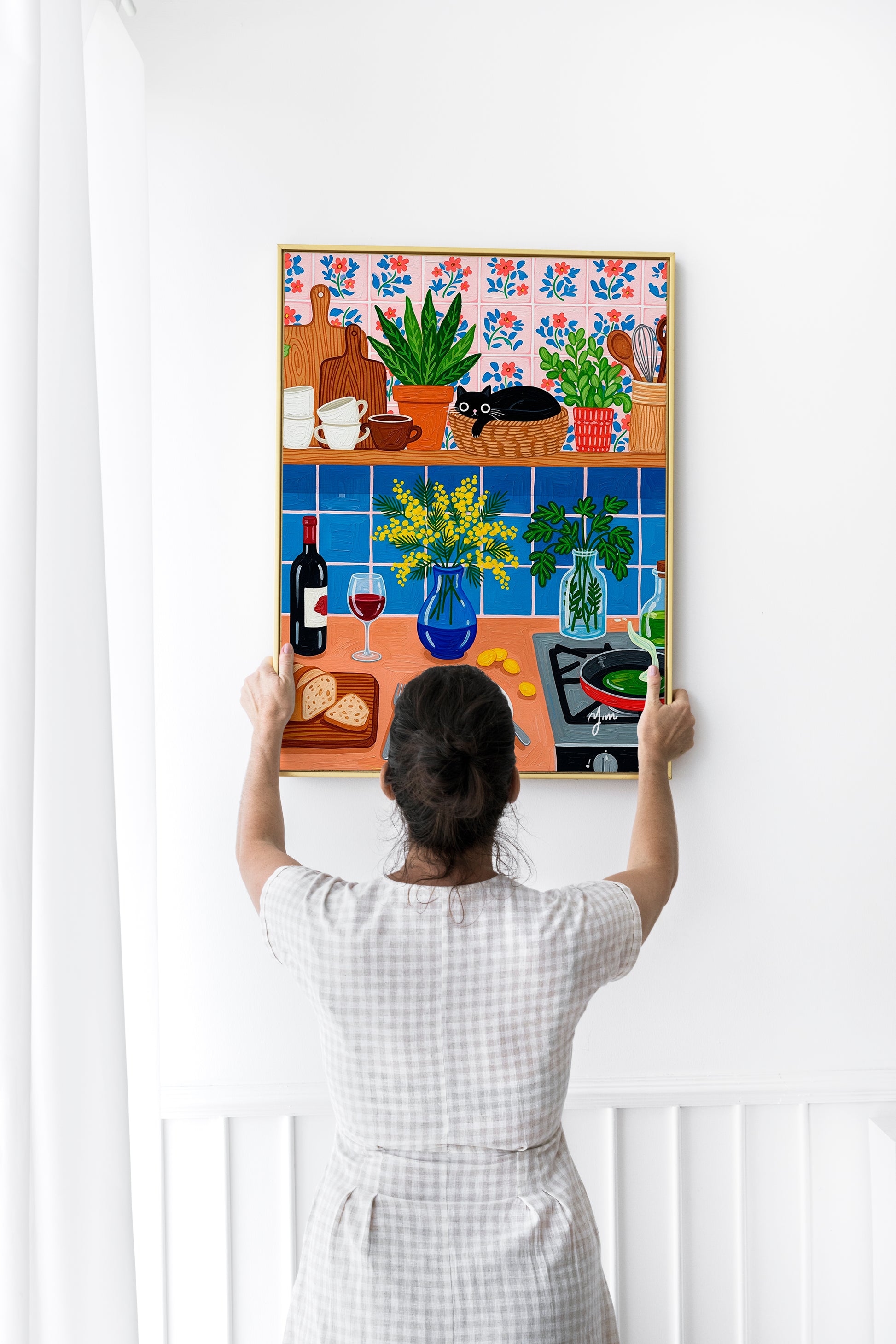 Whimsical kitchen illustration featuring a black cat curled up in a round wicker basket on a wooden shelf lined with stacked mugs, cutting boards, and potted houseplants. The wall is covered in hand‑painted pink tiles with blue and red floral motifs. Below, a blue tiled backsplash frames a cheerful countertop scene: a cobalt vase of yellow mimosa blossoms, fresh parsley in a glass jar, a bottle and glass of red wine, sliced artisan bread on a wooden board, a plate with two sunny‑side‑up eggs, scattered gold