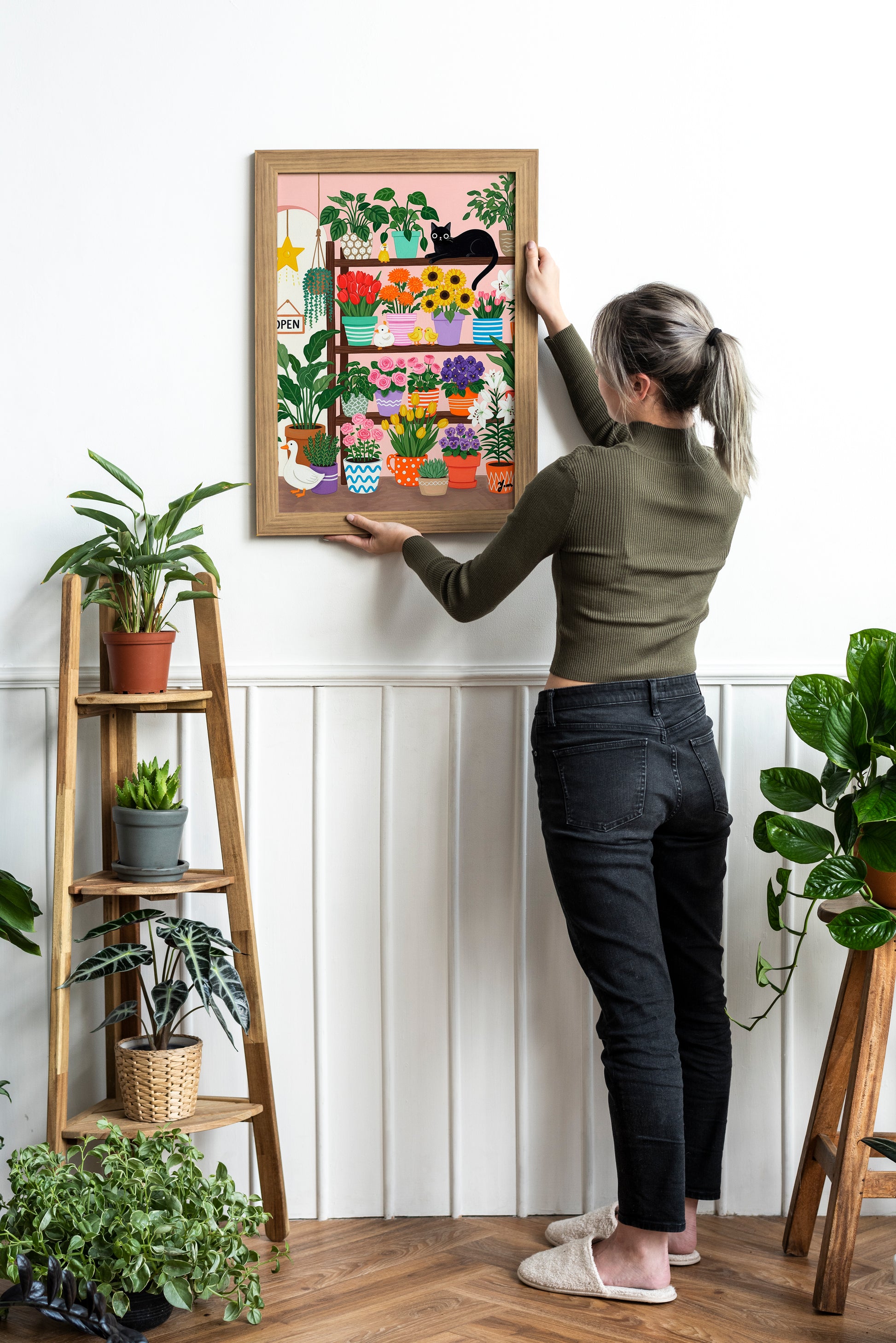 A whimsical illustration of a cozy flower shop filled with colorful potted flowers like tulips, roses, sunflowers, and lilies. A black cat lounges on a shelf, surrounded by cheerful ducks and a white goose. The scene is bright, playful, and perfect for spring or cottagecore decor.